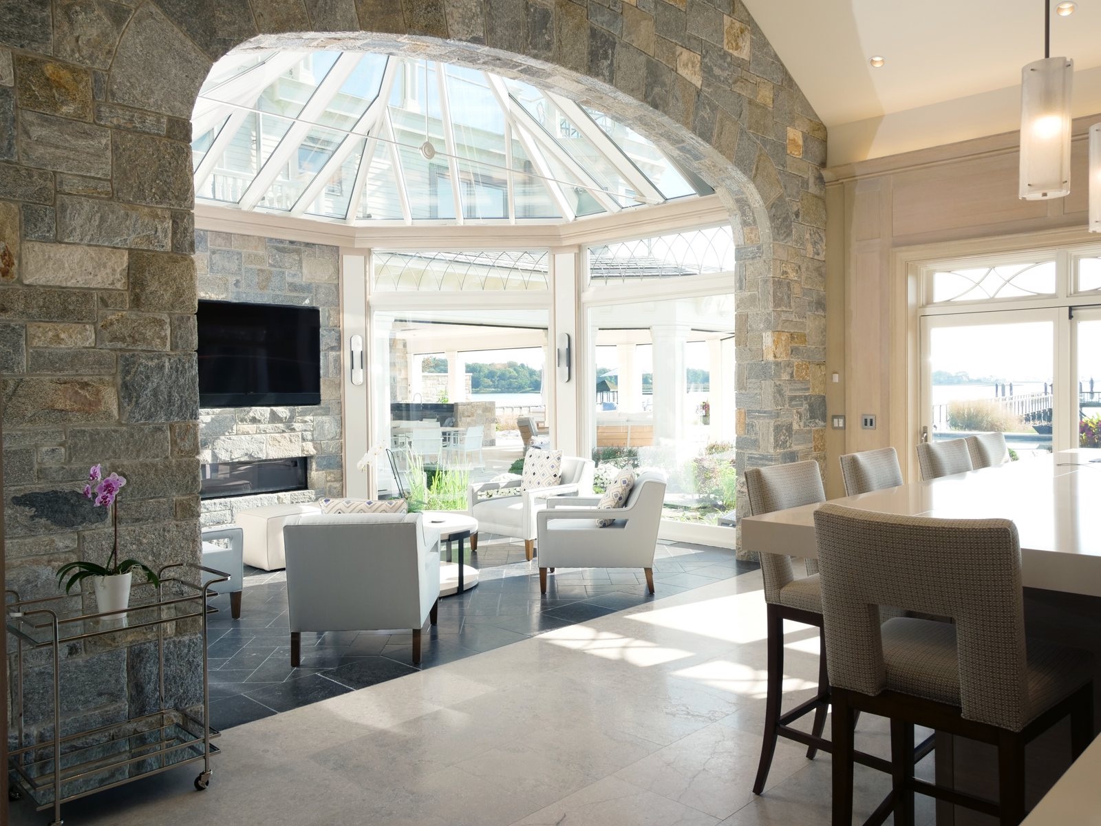 Kitchen with a pale limestone floor opening through a building stone arch into a glass conservatory overlooking the water
